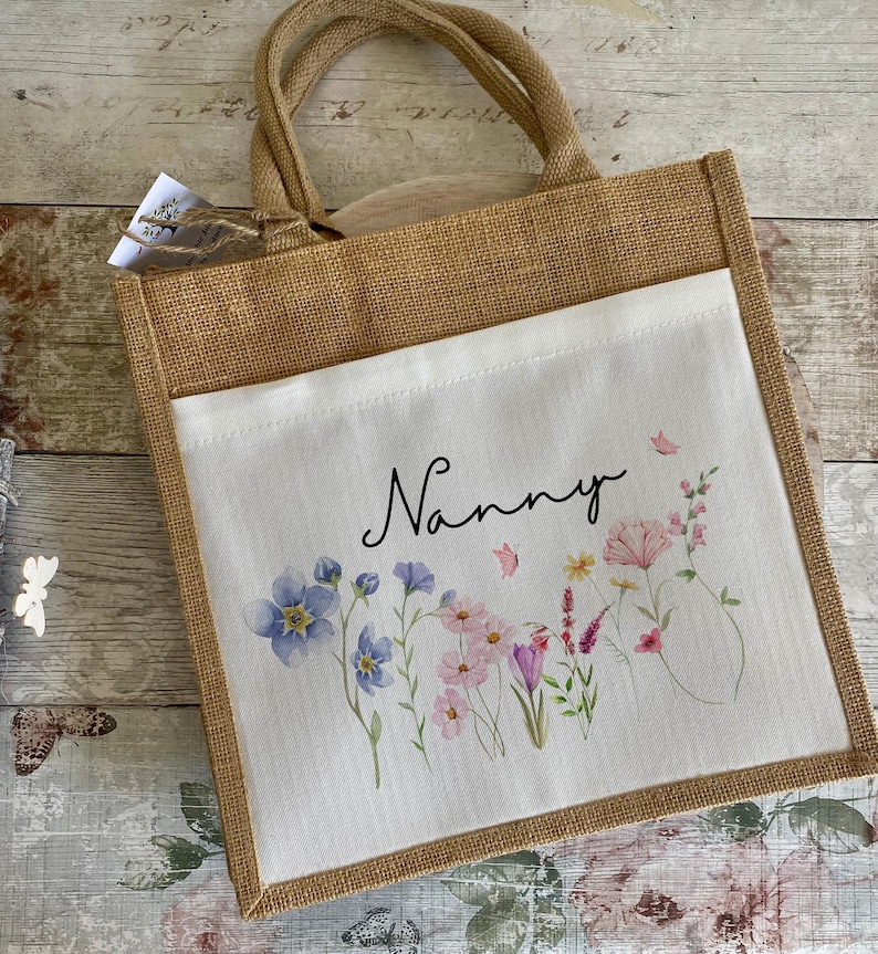 K&ouml;nnte beinhalten: Eine braune Jute-Tragetasche mit einem wei&szlig;en Stoffeinsatz. Der Einsatz zeigt ein Blumenmuster mit rosa, lila und blauen Blumen und dem Wort "Nanny" in schwarzer Schrift.
