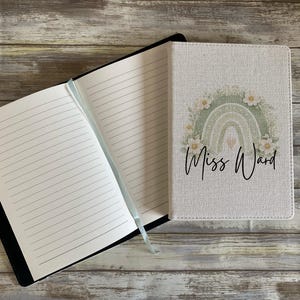 May include: A notebook with a textured off-white cover featuring a sage green rainbow graphic, white flowers, and the text Miss Ward in black script. The notebook is open to reveal lined white pages and a light blue ribbon bookmark attached to the spine.