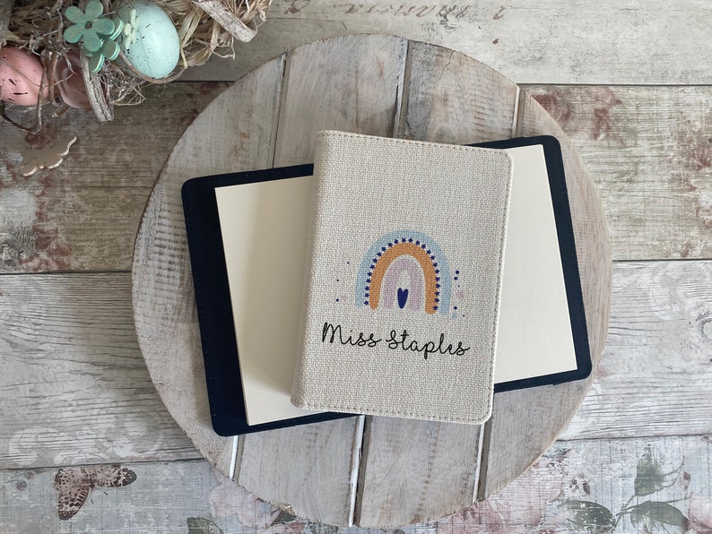 K&ouml;nnte beinhalten: Ein beigefarbener Leinen-Notizbuchumschlag mit einem Regenbogen- und Herzdesign. Der Text "Miss Staplus" ist auf dem Umschlag aufgedruckt.