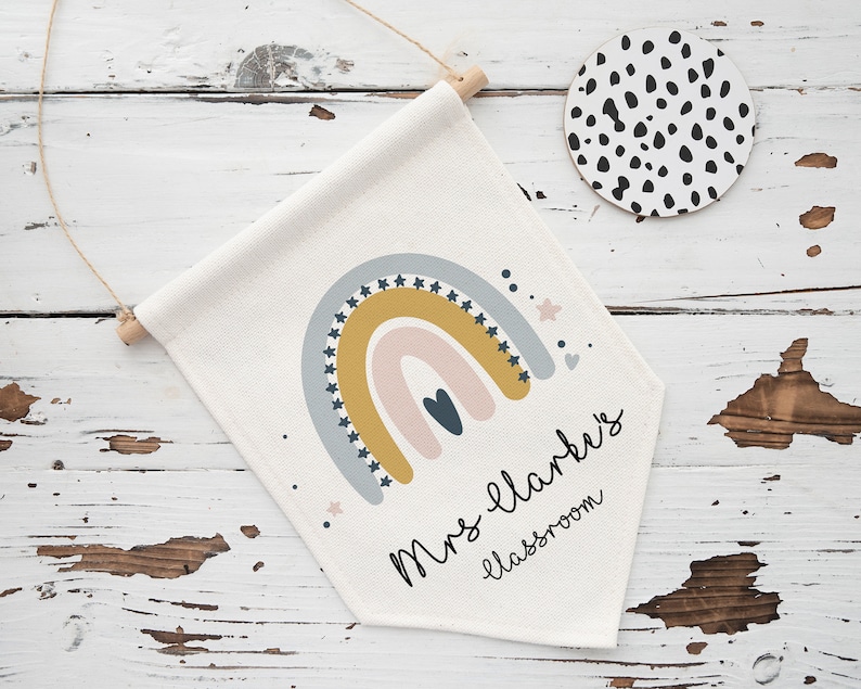 May include: A cream-colored classroom banner with a rainbow design in muted colors, including gold, blue, and pink. The banner has the text "Mrs. Clark's classroom" in a handwritten font. A round coaster with black spots is also visible.