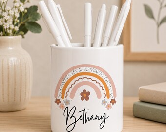 May include: A white ceramic pencil holder featuring a printed design of a multi-colored rainbow in shades of pink, orange, and brown, accented with small flowers and the name Bethany written in black cursive script, containing several white pens.