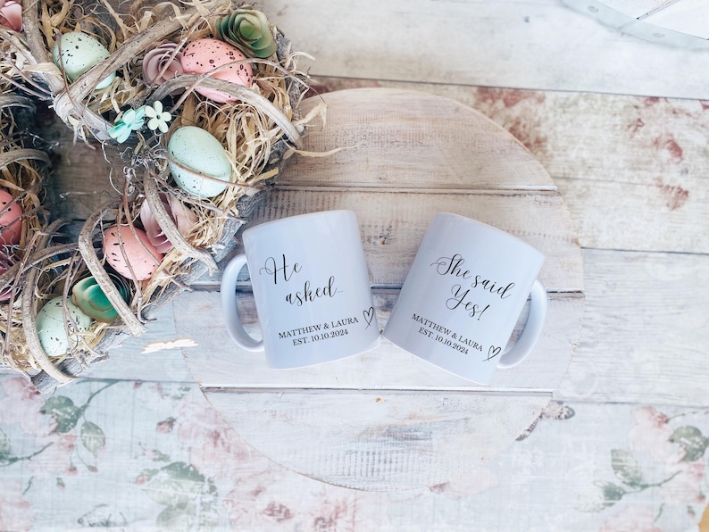 May include: Two white ceramic mugs with the phrases "He asked" and "She said Yes!" in elegant script. Each mug includes the names "Matthew & Laura" and the date "EST. 10.10.2024". A heart symbol is below the names. The mugs are on a light wood surface.