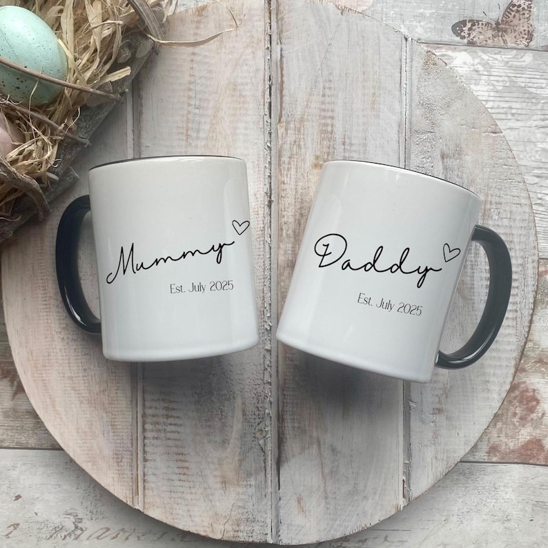 May include: Two white ceramic mugs with black handles and rims. One mug says "Mummy" and the other says "Daddy" in cursive script, each with a heart and the date "Est. July 2025". The mugs are on a wooden surface.
