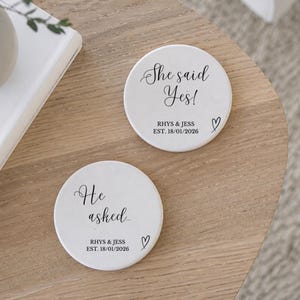 May include: Two white ceramic coasters with black text and a heart symbol. One coaster reads "She said Yes!" with the names "RHYS & JESS" and the date "EST. 18/01/2026". The other coaster reads "He asked..." with the same names and date.