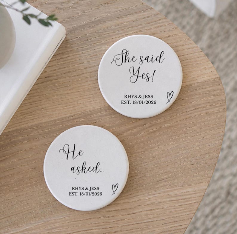 May include: Two white ceramic coasters with black text and a heart symbol. One coaster reads "She said Yes!" with the names "RHYS & JESS" and the date "EST. 18/01/2026". The other coaster reads "He asked..." with the same names and date.
