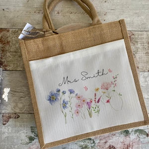 May include: A square jute tote bag with two top handles and a white canvas front pocket. The pocket features a watercolour illustration of various wildflowers in shades of blue, pink, and yellow, with the text Mrs Smith printed in black script.