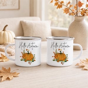 May include: Two white enamel mugs with a silver rim, each featuring a graphic of an orange pumpkin surrounded by autumn leaves and the black cursive text Hello Autumn. The mugs have a side handle and a smooth, glossy finish on the exterior surface.