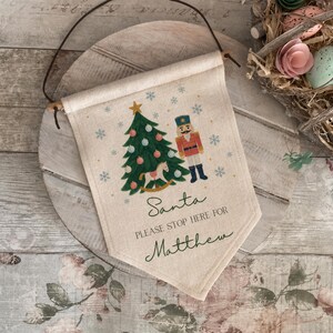 May include: A cream-coloured fabric wall banner featuring a printed Christmas tree, a nutcracker, and a rocking horse. The text reads Santa PLEASE STOP HERE FOR Matthew. The banner is suspended by a dark brown cord attached to a wooden dowel rod.