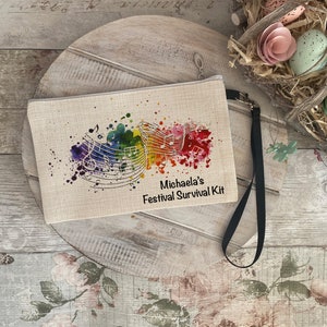 Op de afbeelding: Een beige canvas tasje met een zwarte rits en een zwarte band. Het tasje heeft een kleurrijk aquarel ontwerp met muzieknoten en de tekst "Michaela's Festival Survival Kit".