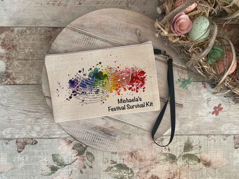 Op de afbeelding: Een beige canvas tasje met een zwarte rits en een zwarte band. Het tasje heeft een kleurrijk aquarel ontwerp met muzieknoten en de tekst "Michaela's Festival Survival Kit".