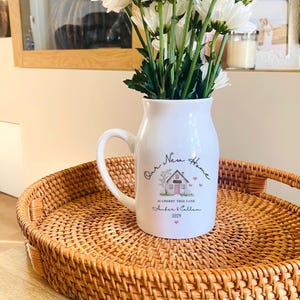 May include: A white ceramic pitcher vase with a handle, filled with white daisies. The front features a printed illustration of a house with a pink door, the text Our New Home, 16 Cherry Tree Lane, Amber & Callam, and the year 2025 with small hearts.