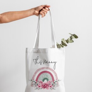 Op de afbeelding: Witte draagtas met de tekst "This Nanny" en een regenboogontwerp in roze, groen en wit. De onderkant van de tas heeft een bloemstuk en de tekst "Belongs To Matthew & Juliette XX".