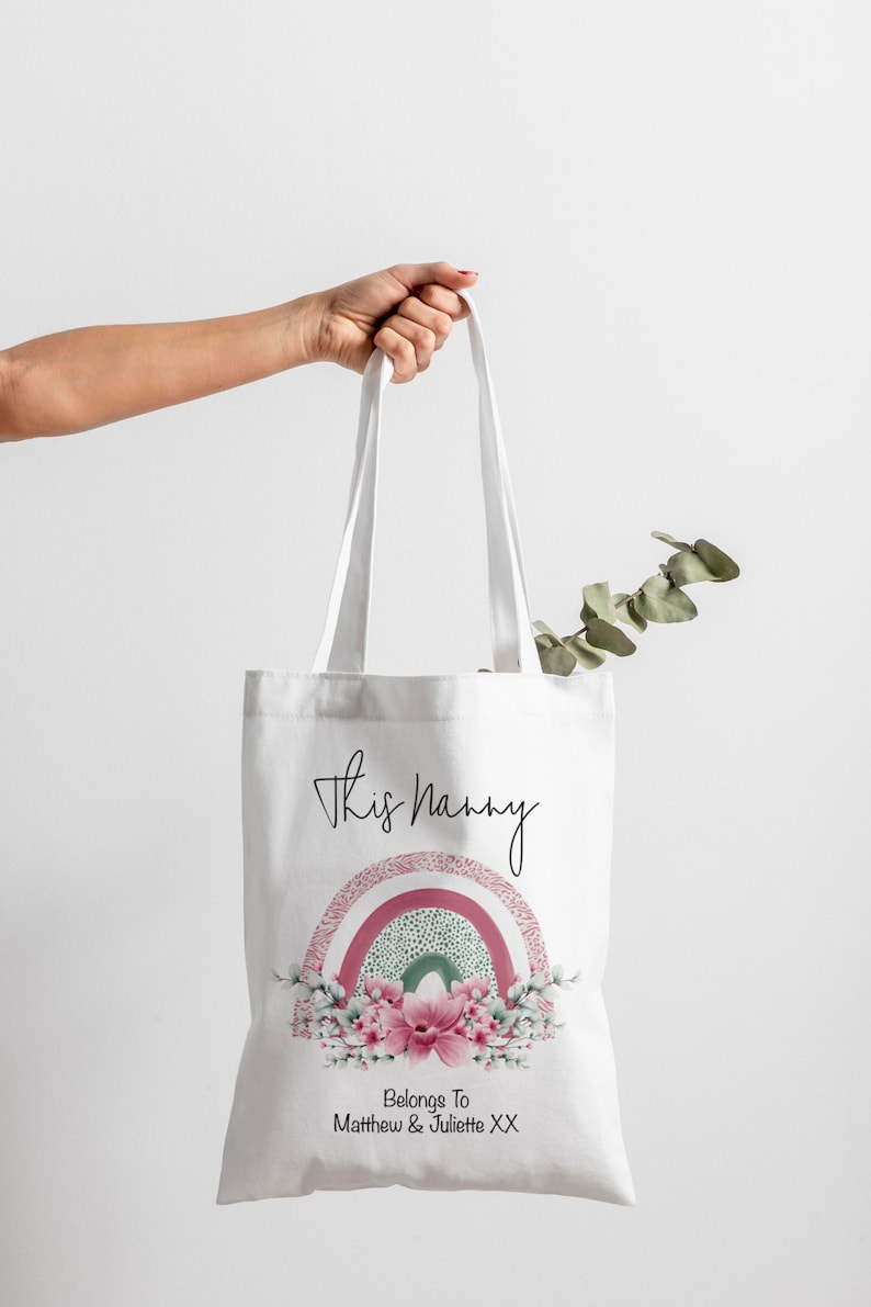 Op de afbeelding: Witte draagtas met de tekst "This Nanny" en een regenboogontwerp in roze, groen en wit. De onderkant van de tas heeft een bloemstuk en de tekst "Belongs To Matthew & Juliette XX".
