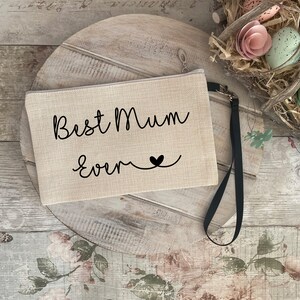 May include: A beige canvas zippered pouch with a black strap. The pouch has the text "Best Mum Ever" in black cursive lettering with a small black heart.