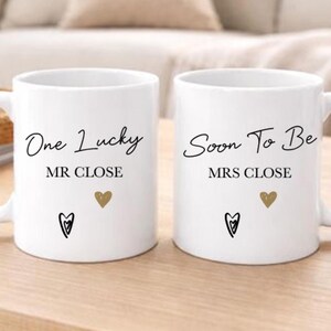 May include: Two white ceramic mugs with black text that reads "One Lucky MR CLOSE" and "Soon To Be MRS CLOSE". Each mug has a gold heart and a black heart design. The mugs are on a light wood surface.