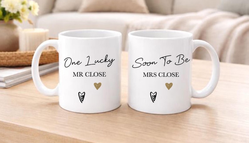 May include: Two white ceramic mugs with black text that reads "One Lucky MR CLOSE" and "Soon To Be MRS CLOSE". Each mug has a gold heart and a black heart design. The mugs are on a light wood surface.