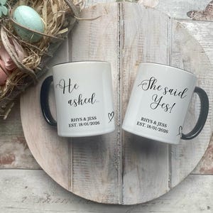 May include: Two white ceramic mugs with black handles and rims. One mug reads "He asked" and the other reads "She said Yes!" in elegant script. Both mugs include the names "RHYS & JESS" and the date "EST. 18/01/2026". A heart symbol is next to the "He asked" mug. The mugs are on a wooden surface.
