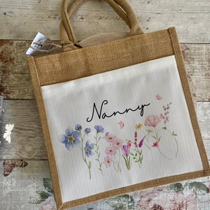 K&ouml;nnte beinhalten: Eine braune Jute-Tragetasche mit einem wei&szlig;en Stoffeinsatz. Der Einsatz zeigt ein Blumenmuster mit rosa, lila und blauen Blumen und dem Wort "Nanny" in schwarzer Schrift.