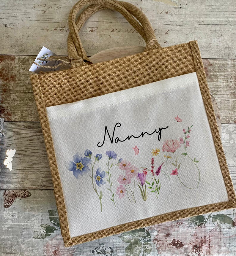 K&ouml;nnte beinhalten: Eine braune Jute-Tragetasche mit einem wei&szlig;en Stoffeinsatz. Der Einsatz zeigt ein Blumenmuster mit rosa, lila und blauen Blumen und dem Wort "Nanny" in schwarzer Schrift.