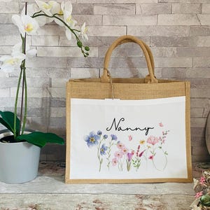 K&ouml;nnte beinhalten: Eine nat&uuml;rliche Jute-Tragetasche mit einem wei&szlig;en Stoffeinsatz. Der Einsatz zeigt ein Blumenmuster mit rosa, blauen und gr&uuml;nen Blumen und dem Wort "Nanny" in schwarzer Schrift.