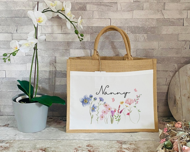 K&ouml;nnte beinhalten: Eine nat&uuml;rliche Jute-Tragetasche mit einem wei&szlig;en Stoffeinsatz. Der Einsatz zeigt ein Blumenmuster mit rosa, blauen und gr&uuml;nen Blumen und dem Wort "Nanny" in schwarzer Schrift.
