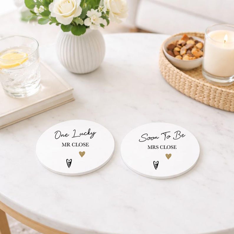 May include: Two white round coasters with black text that reads "One Lucky MR CLOSE" and "Soon To Be MRS CLOSE". Each coaster has a gold heart and a black heart illustration. A vase of white flowers, a candle, and a glass of water are in the background.