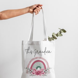 Op de afbeelding: Witte draagtas met de tekst "This grandma". Onder de tekst staat een bloemenregenboogontwerp in roze, groen en wit. De tekst "Belongs To Matthew & Juliette XX" staat onderaan.
