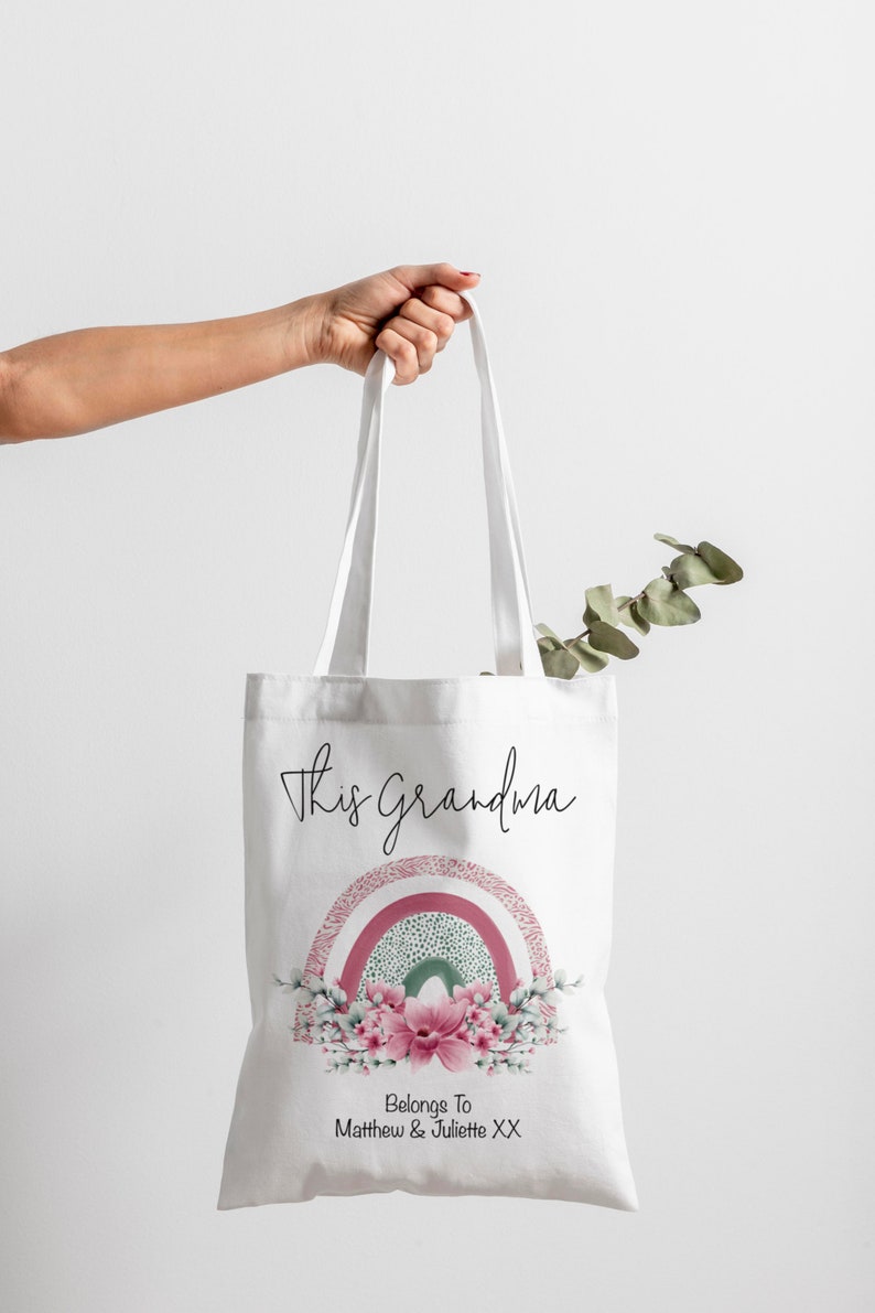Op de afbeelding: Witte draagtas met de tekst "This grandma". Onder de tekst staat een bloemenregenboogontwerp in roze, groen en wit. De tekst "Belongs To Matthew & Juliette XX" staat onderaan.