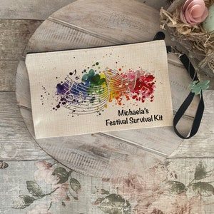 Op de afbeelding: Een witte canvas ritszak met een zwarte band. De tas heeft een kleurrijk aquarelontwerp met muzieknoten en de tekst "Michaela's Festival Survival Kit".