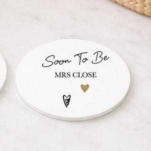 May include: A white, circular coaster with the words "Soon To Be MRS CLOSE" in black script. It features a gold heart and a black heart design. The coaster is likely made of ceramic or a similar material, perfect for a wedding.