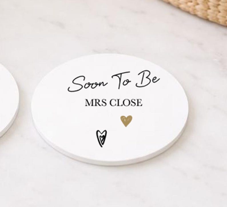 May include: A white, circular coaster with the words "Soon To Be MRS CLOSE" in black script. It features a gold heart and a black heart design. The coaster is likely made of ceramic or a similar material, perfect for a wedding.