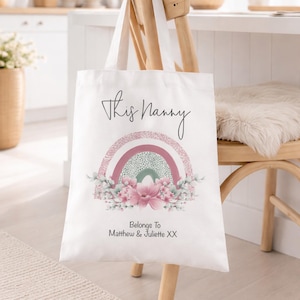 Op de afbeelding: Witte draagtas met de tekst "This Nanny" boven een regenboogontwerp in roze, groen en wit. De onderkant van de tas heeft de tekst "Belongs To Matthew & Juliette XX". De tas hangt aan een houten stoel.