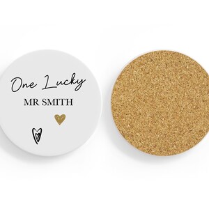 May include: A white ceramic coaster with the words "One Lucky MR SMITH" in black script, a gold heart, and a black heart. The reverse side is made of cork.