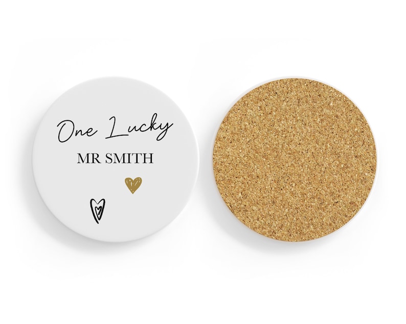 May include: A white ceramic coaster with the words "One Lucky MR SMITH" in black script, a gold heart, and a black heart. The reverse side is made of cork.