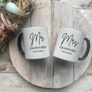 May include: Two white ceramic mugs with black handles and black text. The mug on the left says "Mr. Crawford 06.09.2024" and the mug on the right says "Mrs. Crawford 06.09.2024".
