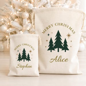 May include: Two off-white drawstring gift bags made of fabric. Each bag features a dark green graphic of three pine trees, gold stars, and the text Merry Christmas. The larger bag is personalized with the name Alice and the smaller with Stephen.