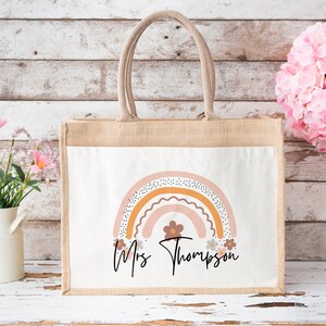 May include: A natural jute tote bag with a white canvas insert featuring a rainbow design with pink, orange, and brown colors. The bag has a personalized message that reads "Mrs. Thompson" with a small flower design.