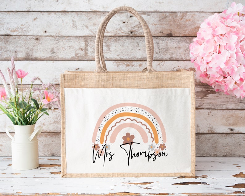 May include: A natural jute tote bag with a white canvas insert featuring a rainbow design with pink, orange, and brown colors. The bag has a personalized message that reads "Mrs. Thompson" with a small flower design.