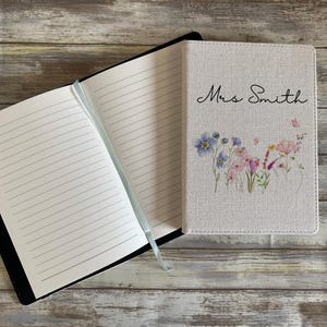 May include: A notebook with a light beige fabric cover featuring a watercolor floral print and the text Mrs. Smith in black script. The notebook is open to reveal white lined pages and includes a light blue ribbon bookmark attached to the spine.
