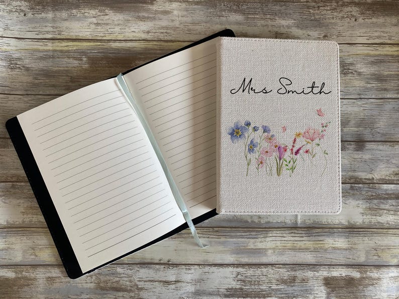 May include: A notebook with a light beige fabric cover featuring a watercolor floral print and the text Mrs. Smith in black script. The notebook is open to reveal white lined pages and includes a light blue ribbon bookmark attached to the spine.