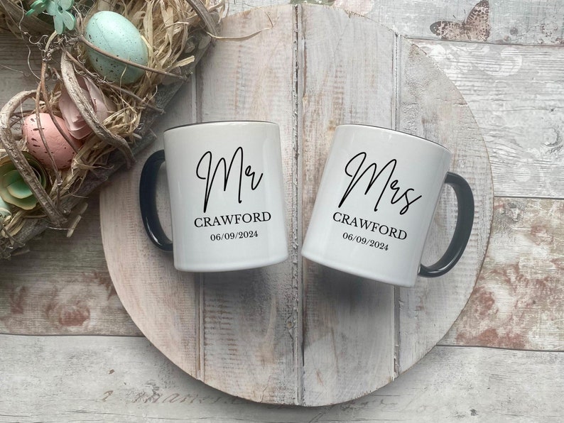 May include: Two white ceramic mugs with black handles and black lettering. The mug on the left says "Mr. Crawford 06/09/2024" and the mug on the right says "Mrs. Crawford 06/09/2024".