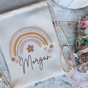 May include: A white drawstring bag with a colorful rainbow design and the name "Morgan" printed below. The rainbow is made up of pink, orange, and yellow stripes with white polka dots. The bag is decorated with small pink and white flowers.