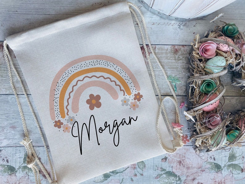 May include: A white drawstring bag with a colorful rainbow design and the name "Morgan" printed below. The rainbow is made up of pink, orange, and yellow stripes with white polka dots. The bag is decorated with small pink and white flowers.