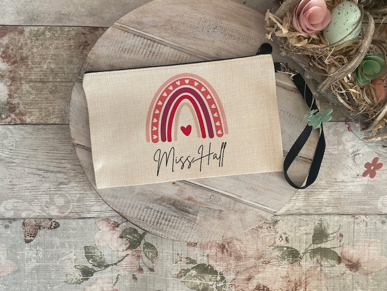 Pode incluir: Uma bolsa de lona bege com z&iacute;per com um design de arco-&iacute;ris e o texto "Miss Hall". O arco-&iacute;ris &eacute; feito de listras rosa, vermelhas e laranja com cora&ccedil;&otilde;es brancos ao redor das bordas. A bolsa tem um z&iacute;per preto e uma al&ccedil;a preta.