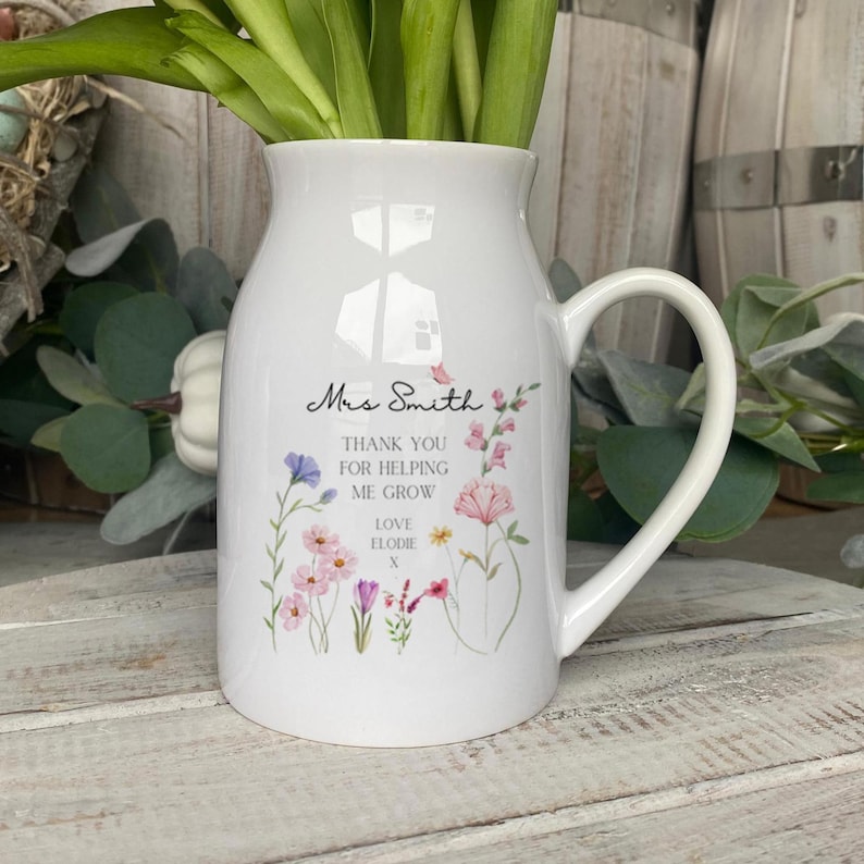 May include: A white ceramic pitcher vase with a handle, featuring a floral print and black text that reads: Mrs Smith, THANK YOU FOR HELPING ME GROW, LOVE ELODIE x. The design includes various colourful wildflowers in shades of pink, purple, and yellow.