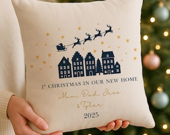 May include: A beige throw pillow with a Christmas theme. It features a navy blue illustration of Santa's sleigh and houses, with gold stars. Text reads "1st Christmas in Our New Home" and names, with the year 2025.