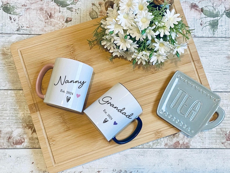 Op de afbeelding: Twee witte keramische mokken met zwarte letters. De mok aan de linkerkant zegt "Nanny Est. 2024" met een roze hart. De mok aan de rechterkant zegt "Grandad Est. 2024" met een paars hart. Een grijze keramische theekopschotel met het woord "TEA" in witte letters is op de achtergrond.