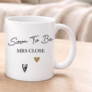 May include: White ceramic mug with the text "Soon To Be MRS CLOSE" in black script. The mug features a gold heart and a black heart design. The mug has a curved handle and is likely used for beverages.
