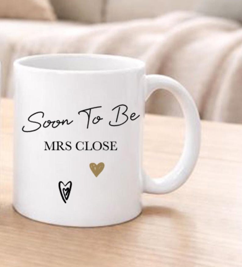 May include: White ceramic mug with the text "Soon To Be MRS CLOSE" in black script. The mug features a gold heart and a black heart design. The mug has a curved handle and is likely used for beverages.