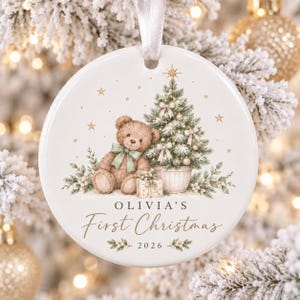 May include: A round white ceramic Christmas ornament featuring a brown teddy bear with a green bow, a decorated Christmas tree, and a gift box. The text reads OLIVIA'S First Christmas 2026, accented with gold stars and greenery.